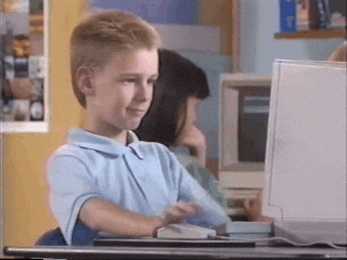 A white boy with blonde hair and a baby blue shirt sits at a computer and gives a thumbs up.
