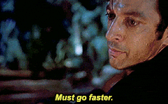 A man with black hair and a black shirt turning to the camera and mouthing, "Must go faster."