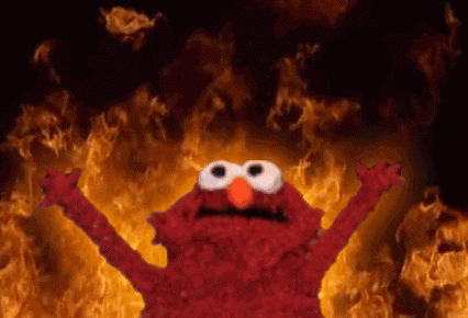 Elmo, a red puppet, standing in front of flames with his arms up.