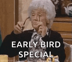 An elderly woman wearing glasses is sitting at a table with a cup of coffee and says "early bird special"