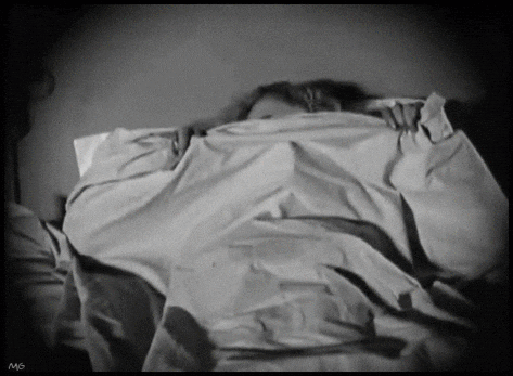 A black and white movie showing a woman fearfully peeping over the covers