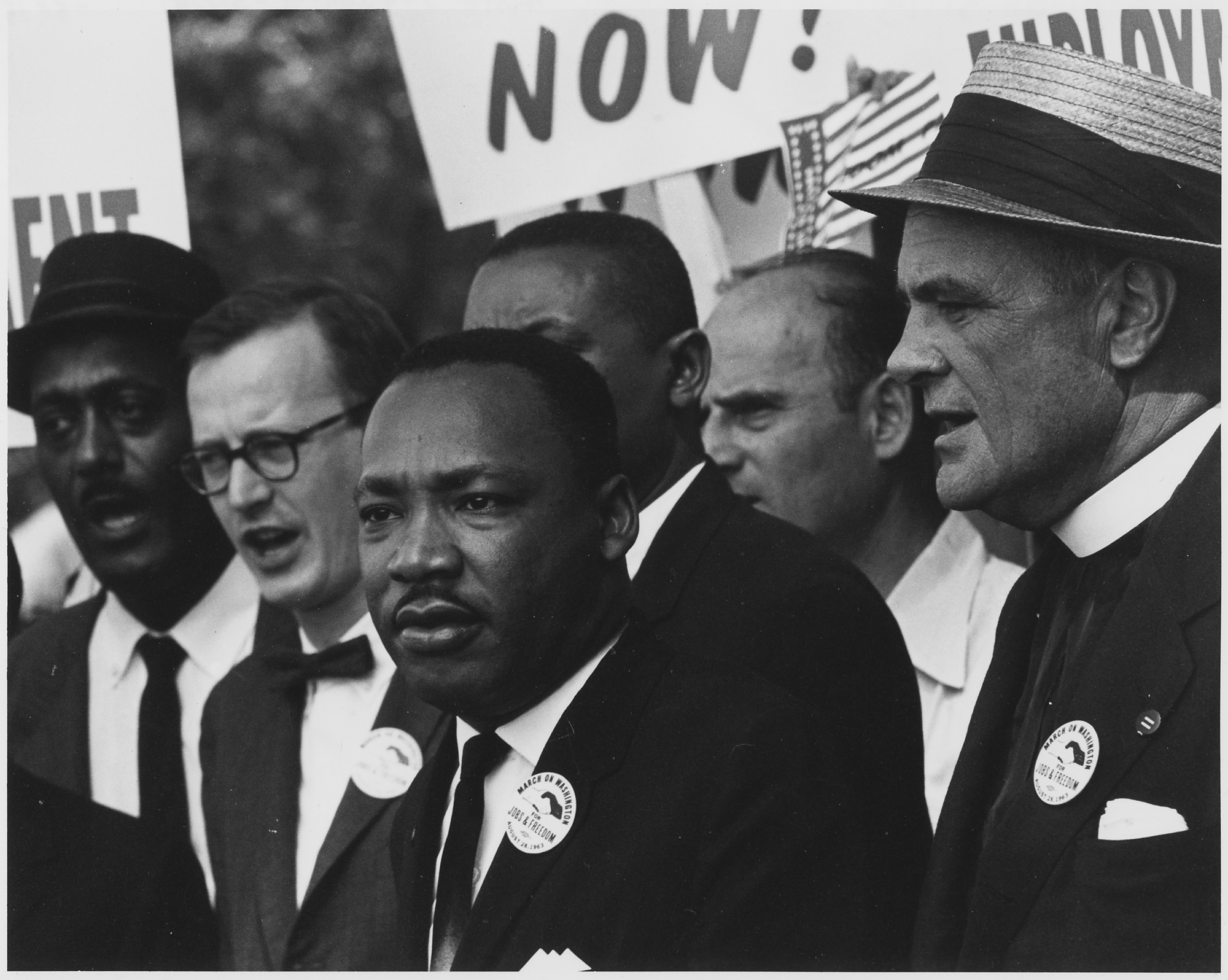 Dr. Martin Luther King, Jr. and Mathew Ahmann in a crowd. There are people holding protest signs in the background.