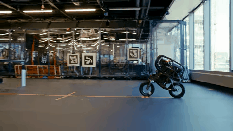 An animated GIF of a robotic bicycle cycling up towards what looks like a ping-pong table, and then leaping up onto it.