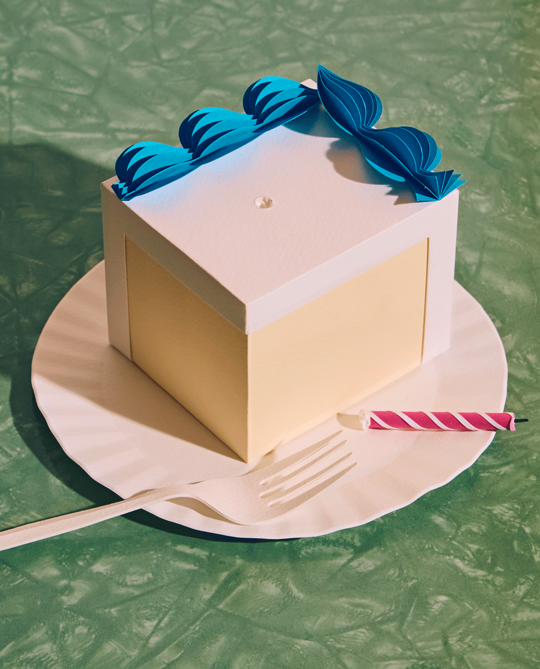 A motion GIF of a square cake piece with blue icing on a paper plate with a pink candle rolling back and forth