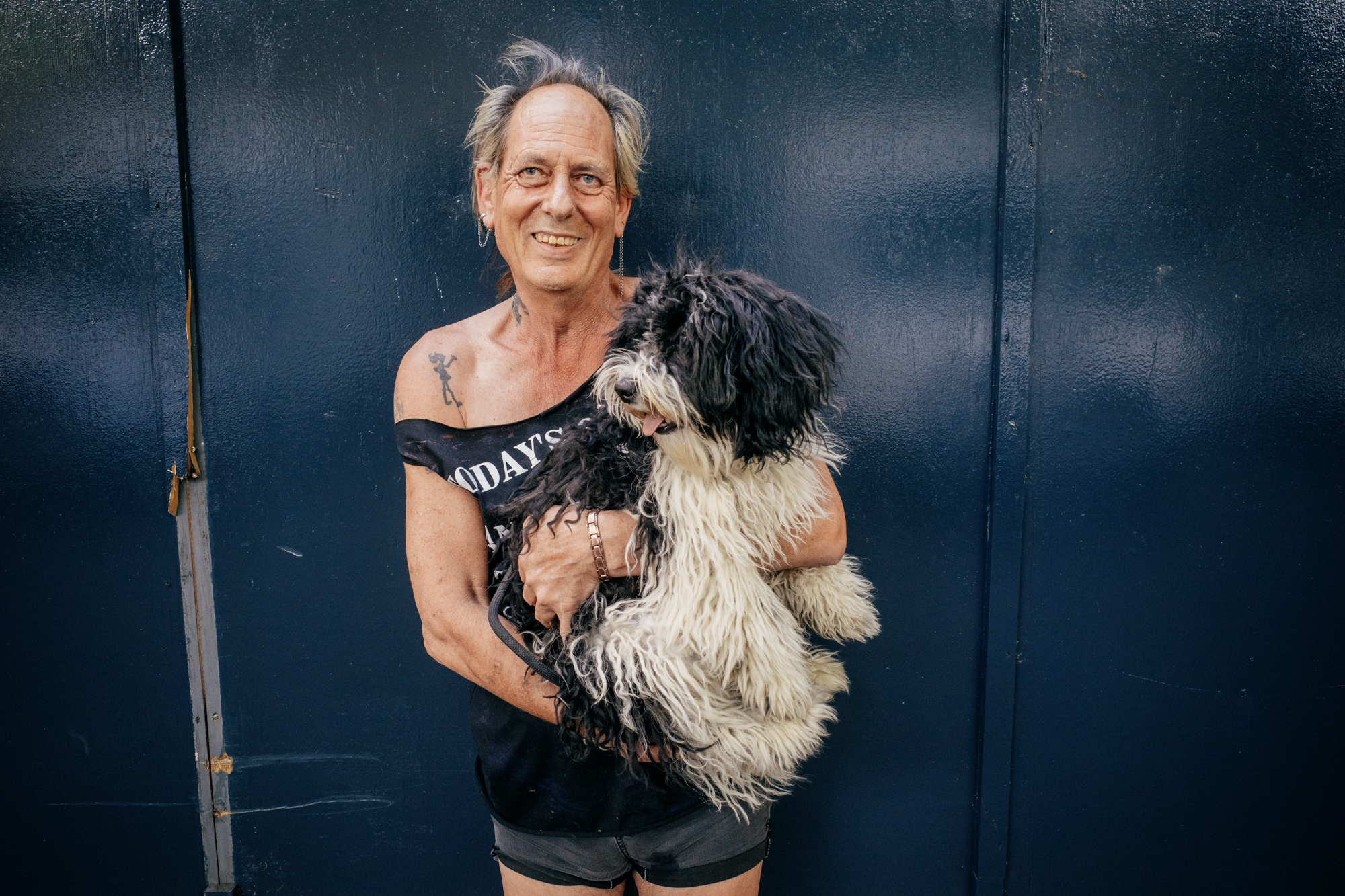 An older punk-looking man standing holding a fluffy black and white dog. He's wearing a black tee with the sleeves cut off and denim shorts