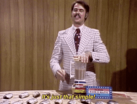 A white man with brown hair in a 70s-style white suit with red accents showcasing a blender and saying, 'It's just that simple!"