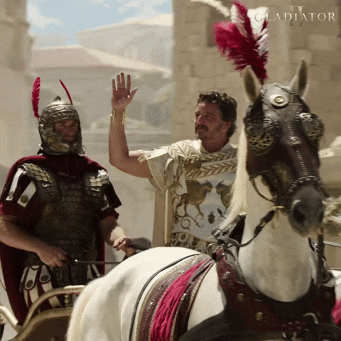 An attractive Roman general in a white tunic and with one hand raised is driven on a chariot through Rome as people on the street cheer him.